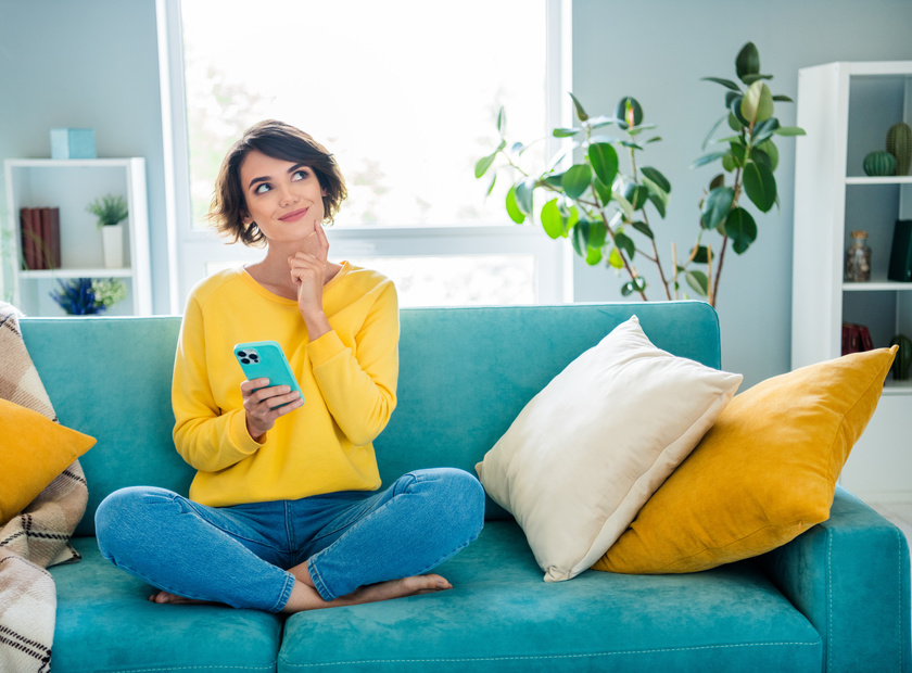 Portrait of pretty dreamy girl sitting on soft cozy couch use phone watching up planning weekend indoor house modern flat.