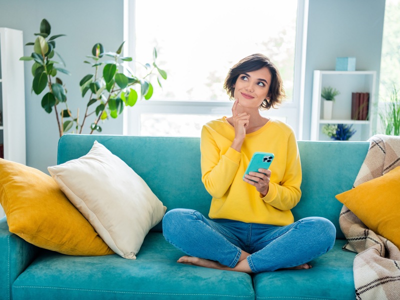 Portrait of pretty dreamy girl sitting on soft cozy couch use phone watching up planning weekend indoor house modern flat.