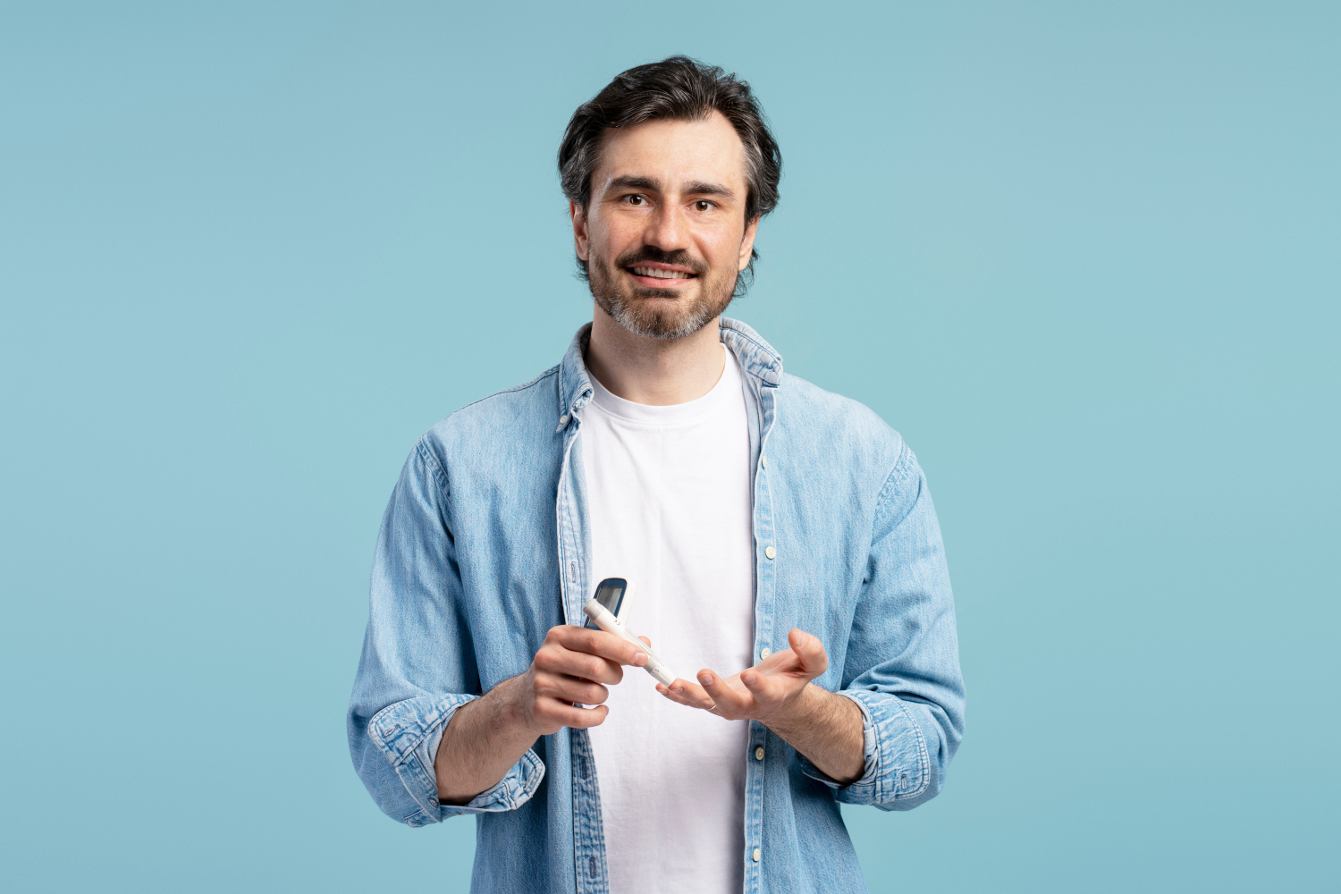 Caucasian man performing diabetes test using glucometer, checking glucose level in blood, smiling and looking at camera on a blue background. Health care concept