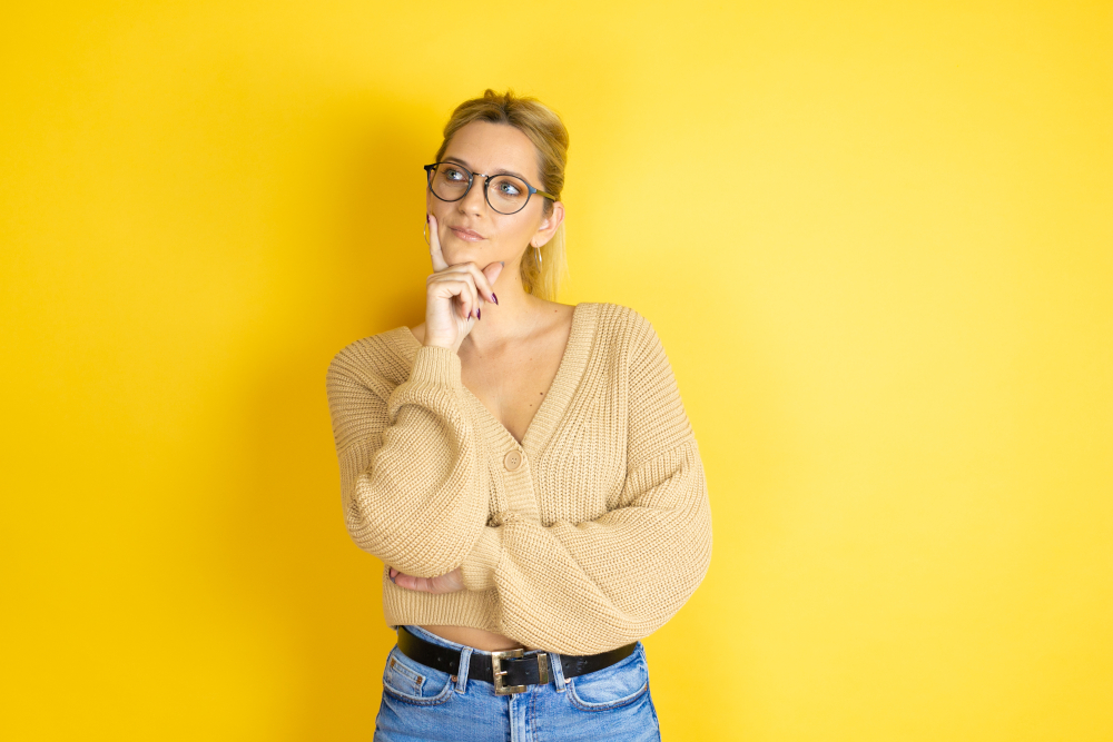 Young beautiful woman wearing casual sweater over isolated yellow background serious face thinking about question with hand on chin, thoughtful about confusing idea